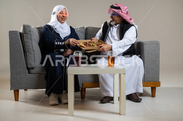 أجواء عائليه سعيده، الاستعداد لتناول الطعام مع الاهل، بورتريه لعائله عربيه خليجيه سعوديه كبار بالسن يجلسان على كنبه مريحه يتناولان طعام العشاء في غرفه المعيشه، رجل وأمراه  يرتديان الزي السعودي التقليدي ، مأكولات ومشروبات سريعه وجاهزه، خلفيه رماديه