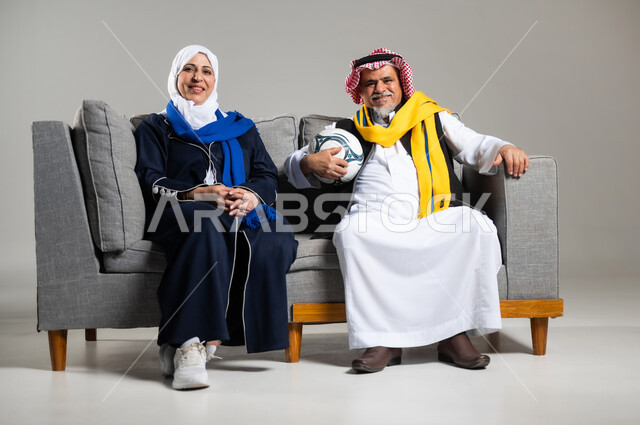 الشغف بمباريات كرة القدم ، قضاء وقت ممتع بمشاهدة ومتابعة مباريات كرة القدم،  بورتريه لزوجان عربيان خليجيان سعوديان كبار بالسن يجلسون على كنبه مريحه في غرفه المعيشه ينظران الى التلفاز بتركيز وتمعن، ارتداء أوشحه تشجيعيه لفريق كره القدم ، خلفية رماديه