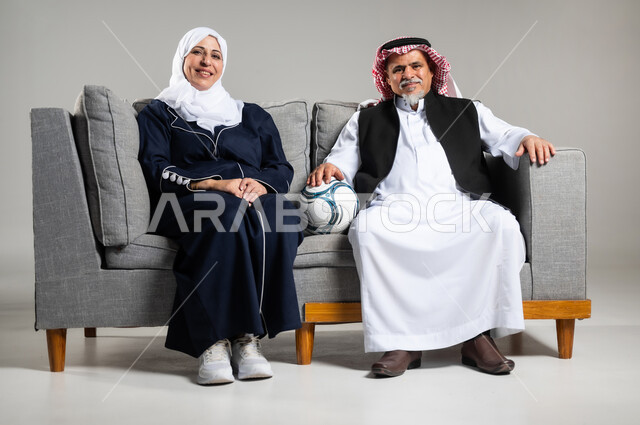 الشغف بمباريات كرة القدم ، قضاء وقت ممتع بمشاهدة ومتابعة مباريات كرة القدم،  بورتريه لزوجان عربيان خليجيان سعوديان كبار بالسن يجلسون على كنبه مريحه في غرفه المعيشه ينظران الى التلفاز بتركيز وتمعن، ارتداء أوشحه تشجيعيه لفريق كره القدم ، خلفية رماديه