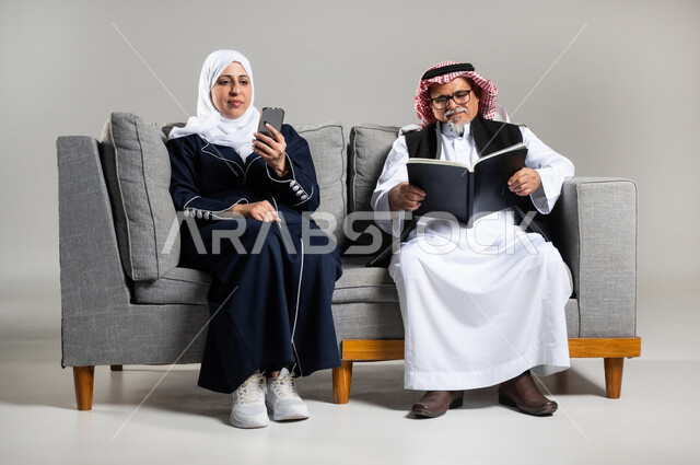 التزود بالعلم والمعرفه، قضاء وقت الفراغ بالتثقيف والتعلم، مفهوم محو الاميه، بورتريه لرجل عربي خليجي سعودي كبير بالسن يرتدي الزي السعودي والستره الوقائيه يمسك بيده كتابا يقوم بالقراءه يجلس على كنبه مريحه وبجانبه زوجته تمسك بيدها هاتفها المحمول، خلفيه رماديه