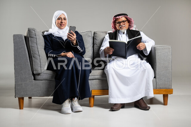 التزود بالعلم والمعرفه، قضاء وقت الفراغ بالتثقيف والتعلم، مفهوم محو الاميه، بورتريه لرجل عربي خليجي سعودي كبير بالسن يرتدي الزي السعودي والستره الوقائيه يمسك بيده كتابا يقوم بالقراءه يجلس على كنبه مريحه وبجانبه زوجته تمسك بيدها هاتفها المحمول، خلفيه رماديه