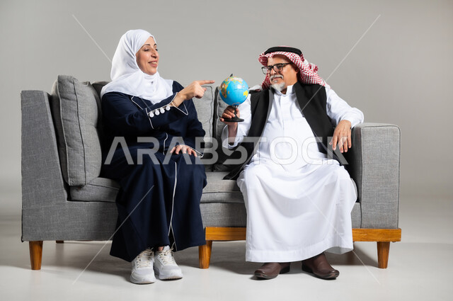 مفهوم البحث والاستكشاف حول العالم, الاشارة إلى مكان معين والنظر إلى أحد القارات, بورتريه لرجل وأمرأه عربيان خليجيان سعوديان كبار بالسن يرتديان الزي السعودي التقليدي  يجلسان على كنبه مريحه  يمسك الرجل بيده مجسما للكره الارضيه وتشير زوجته الى بلد ما، خلفيه رماديه