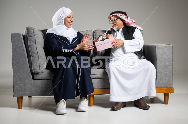 مفهوم الألفة والمحبة بين الزوجين, مشاركة وتقديم الهدايا في الأعياد والمناسبات العائلية, رجل عربي خليجي سعودي كبير بالسن يرتدي الزي السعودي والستره الوقائيه  يقدم لزوجته هدية بمناسبة ذكرى يوم الزواج, رجل وإمرأة سعوديان يحتفلان بعيد الحب في المنزل يجلسان على كنبه مريحه، خلفيه رماديه