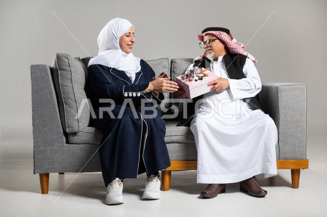 مفهوم الألفة والمحبة بين الزوجين, مشاركة وتقديم الهدايا في الأعياد والمناسبات العائلية, رجل عربي خليجي سعودي كبير بالسن يرتدي الزي السعودي والستره الوقائيه  يقدم لزوجته هدية بمناسبة ذكرى يوم الزواج, رجل وإمرأة سعوديان يحتفلان بعيد الحب في المنزل يجلسان على كنبه مريحه، خلفيه رماديه