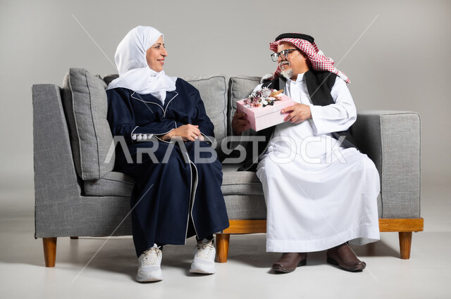 The concept of intimacy and love between spouses, sharing and giving gifts on holidays and family occasions, an elderly Saudi Arabian Gulf man wearing Saudi clothing and a protective vest giving his wife a gift on the occasion of their wedding anniversary, a Saudi man and woman celebrating Valentine's Day at home sitting on a comfortable sofa, gray background