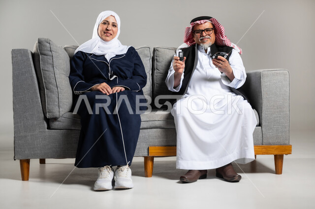 مفهوم القياده في السعوديه , ايماءات الفرح والسرور بامتلاك سيارة جديده, بورتريه لرجل عربي خليجي سعودي مبتسم كبير بالسن يرتدي الزي السعودي والستره الوقائيه يمسك مفتاح التحكم عن بعد ومجسما لسياره صغيره يجلس بجوار زوجته, خلفيه رماديه