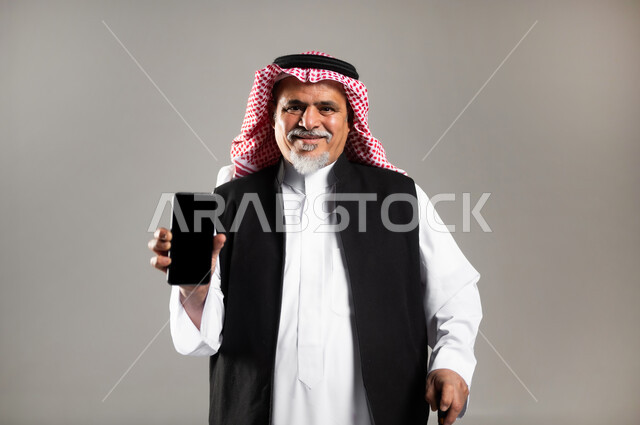 عرض تطبيقات الجوال، بورتريه لرجل عربي خليجي سعودي كبير بالسن يرتدي الزي السعودي والستره الوقائيه يشير بهاتفه المحمول بشاشه سوداء فارغه، التعرف على العروض من خلال الاونلاين، خلفية رماديه