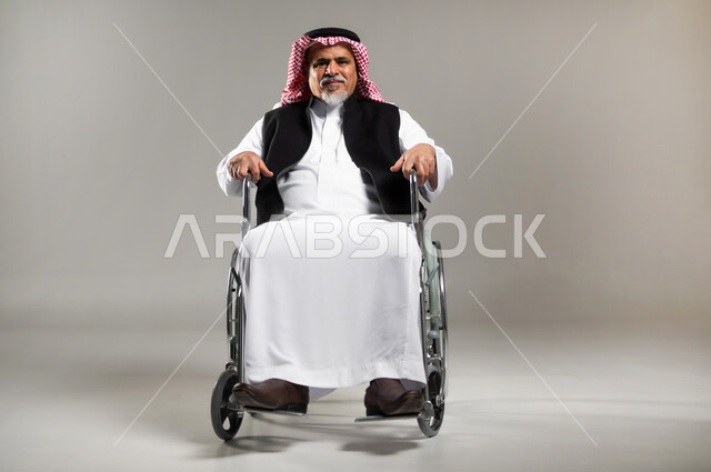 استخدام كراسي المعاقين للمساعده على الحركه والتنقل, تحسين جودة حياة ذوي الاعاقه الحركيه, بورتريه مقرب  لرجل عربي خليجي سعودي كبير بالسن يرتدي الزي السعودي والستره الوقائيه مقعدعلى كرسي متحرك ينظر الى الكاميرا، خلفيه رماديه