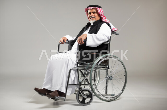 استخدام كراسي المعاقين للمساعده على الحركه والتنقل, تحسين جودة حياة ذوي الاعاقه الحركيه, بورتريه مقرب جانبي لرجل عربي خليجي سعودي كبير بالسن يرتدي الزي السعودي والستره الوقائيه مقعدعلى كرسي متحرك ينظر الى الكاميرا، خلفيه رماديه