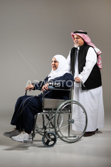 العطف والحنان بين الأزواج، الاهتمام بالجانب النفسي والجسدي للمقعدين، بورتريه لزوج عربي خليجي سعودي كبير بالسن يرتدي الزي السعودي والستره الوقائيه يقوم بجر كرسي زوجته من ذوي الاحتياجات الخاصه، مفهوم  العنايه ورعاية المرضى والمعاقين، ايماءات البشاشه والمعامله الحسنه، خلفيه رماديه