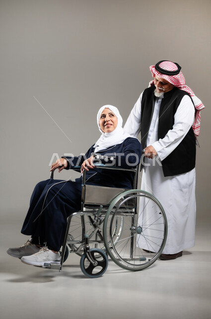 العطف والحنان بين الأزواج، الاهتمام بالجانب النفسي والجسدي للمقعدين، بورتريه لزوج عربي خليجي سعودي كبير بالسن يرتدي الزي السعودي والستره الوقائيه يقوم بجر كرسي زوجته من ذوي الاحتياجات الخاصه، مفهوم  العنايه ورعاية المرضى والمعاقين، ايماءات البشاشه والمعامله الحسنه، خلفيه رماديه