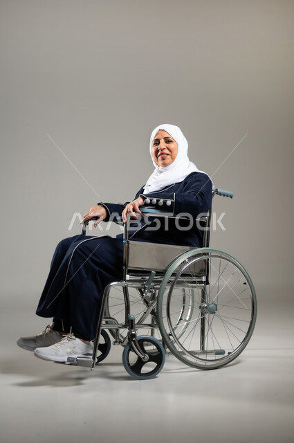 استخدام كراسي المعاقين للمساعده على الحركه والتنقل, تحسين جودة حياة ذوي الاعاقه الحركيه, بورتريه لسيده عربيه خليجيه سعوديه كبيره بالسن ترتدي الحجاب والعباءه السوداء مقعده على كرسي متحرك تنظر الى الكاميرا ببشاشه وابتسامه، خلفيه رماديه