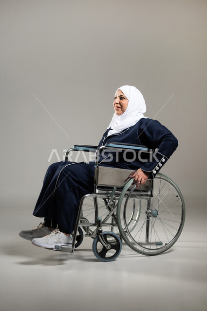 الحفاظ على الصحة البدنية من خلال تحريك اليدين أثناء الحركة, تحسين حياة ذوي الاعاقه الحركيه للمساعده على التنقل, بورتريه لسيده عربيه خليجيه سعوديه محجبه كبيره بالسن ترتدي العباءه السوداء تجلس على كرسي متحرك من ذوي أصحاب الهمم تقوم بتحريكه بنفسها، خلفيه رماديه