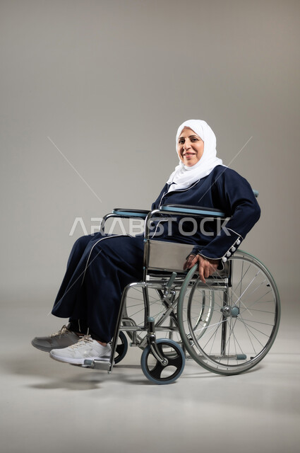 الحفاظ على الصحة البدنية من خلال تحريك اليدين أثناء الحركة, تحسين حياة ذوي الاعاقه الحركيه للمساعده على التنقل, بورتريه لسيده عربيه خليجيه سعوديه محجبه كبيره بالسن ترتدي العباءه السوداء تجلس على كرسي متحرك من ذوي أصحاب الهمم تقوم بتحريكه بنفسها، خلفيه رماديه