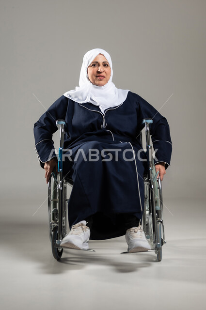 الحفاظ على الصحة البدنية من خلال تحريك اليدين أثناء الحركة, تحسين حياة ذوي الاعاقه الحركيه للمساعده على التنقل, بورتريه لسيده عربيه خليجيه سعوديه محجبه كبيره بالسن ترتدي العباءه السوداء تجلس على كرسي متحرك من ذوي أصحاب الهمم تقوم بتحريكه بنفسها، خلفيه رماديه