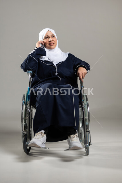 توفير اجهزة ومعدات تساعد بالحركة والتنقل، العنايه بأصحاب الهمم وتأمين احتياجاتهم، بورتريه لسيده عربية سعودية خليجية كبيره بالسن ترتدي الحجاب والعباءة السوداء من ذوي الاحتياجات الخاصه تجلس على كرسي متحرك وتقوم باجراء مكالمه، خلفيه رمادية