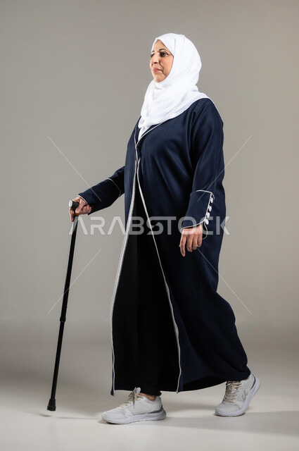 Quality of life of the elderly in Saudi Arabia, walking and mobility with the help of a medical crutch, portrait of an elderly veiled Saudi Arabian Gulf woman wearing a black abaya walking with a crutch in her hand, aging and the need to use assistive devices, gray background
