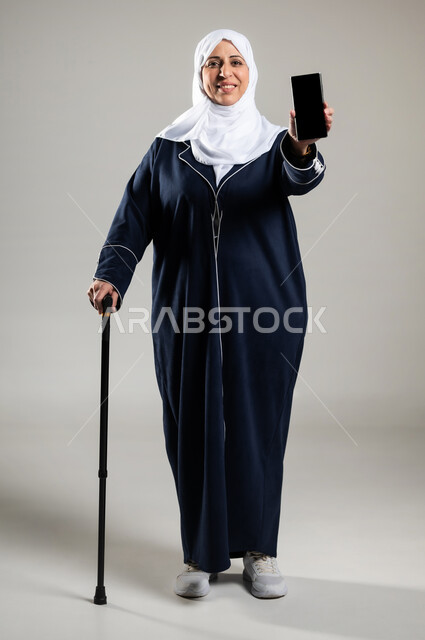 التسويق الالكتروني باستخدام الهاتف المحمول، عرض تطبيقات الاون لاين، بورتريه لسيده عربيه خليجيه سعوديه محجبه ترتدي الحجاب والعباءة السوداء تستند على عكاز وتشير بشاشة سوداء فارغة، الوقوف باستقامه والنظرالى الكاميرا بابتسامه، خلفية رماديه