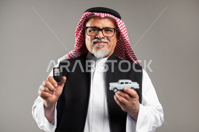 مفهوم القياده في السعوديه، الفرح والسرور بامتلاك سياره حديثه، بورتريه مقرب لرجل عربي خليجي سعودي كبير بالسن يرتدي الثوب التقليدي والشماغ السعودي والستره السوداء الوقائيه يمسك بيده مجسما لسياره صغيره ومفتاح القياده ينظر الى الكاميرا بابتسامه، خلفية رمادية