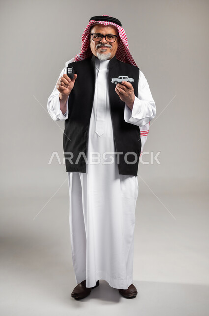 مفهوم القياده في السعوديه، الفرح والسرور بامتلاك سياره حديثه، بورتريه  لرجل عربي خليجي سعودي كبير بالسن يرتدي الثوب التقليدي والشماغ السعودي والستره السوداء الوقائيه يمسك بيده مجسما لسياره صغيره ومفتاح القياده، الوقوف باستقامه والنظر الى الكاميرا بابتسامه، صوره الجسم بالطول الكامل، خلفية رمادية