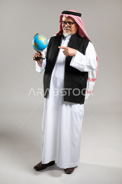 The concept of research and exploration, pointing to a place on the map and looking at one of the continents, a portrait of an old Saudi Gulf Arab man wearing the traditional thobe, the Saudi shemagh, and a black protective jacket, holding a model of the globe in his hand and pointing to it, full-length body image, gray background.