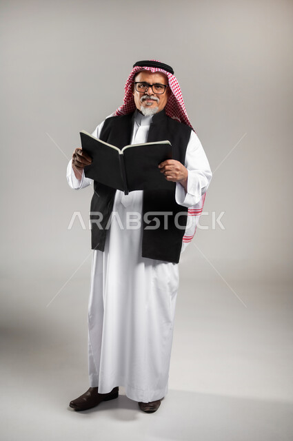 التزود بالعلم والمعرفه، قضاء وقت الفراغ بالتثقيف والتعلم، مفهوم محو الاميه، بورتريه لرجل عربي خليجي سعودي كبير بالسن يرتدي الثوب التقليدي والشماغ السعودي والستره السوداء الوقائيه يحمل بيده كتابا يقوم بالقراءه, الوقوف باستقامة والنظر إلى الكاميرا بإيماءات السرور والبشاشة, صورة الجسم بالطول الكامل, خلفية رمادية