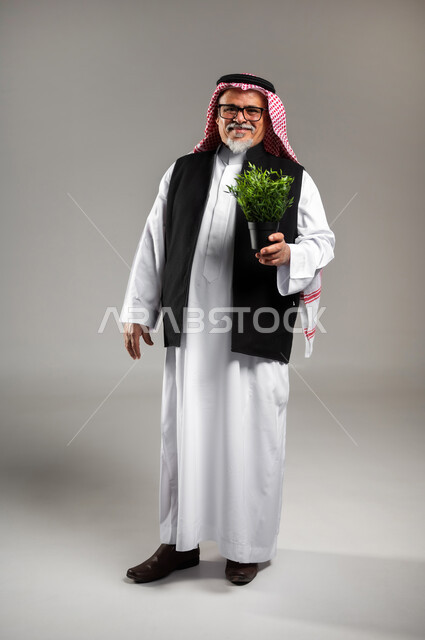 Purifying the air through ornamental plants, taking care of household seedlings, a portrait of an elderly Saudi Arabian Gulf man wearing the traditional thobe, the Saudi shemagh, and a black protective jacket, holding a small agricultural basin in his hand, standing straight and looking at the camera with gestures of pleasure and joy, full-length body portrait, gray background