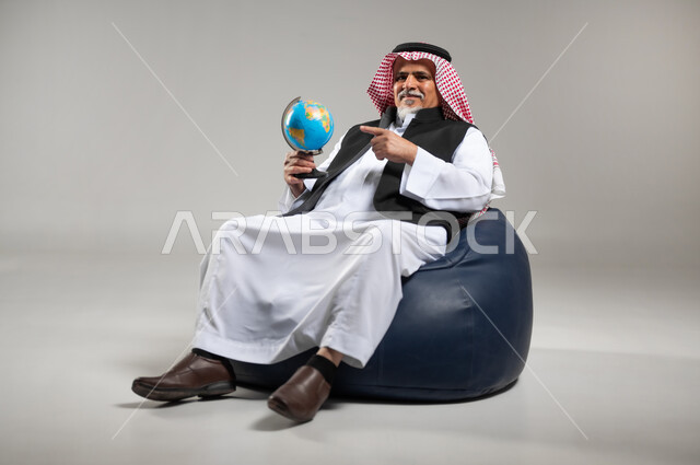 مفهوم البحث والاستكشاف حول العالم, الاشارة إلى مكان معين على الخريطة والنظر إلى أحد القارات, بورتريه لرجل عربي خليجي سعودي كبير بالسن يرتدي الزي السعودي التقليدي والستره السوداء  يجلس على اريكه مريحه من الجلد الفاخر يمسك بيده مجسما للكره الارضيه ويشير اليها، خلفيه رماديه