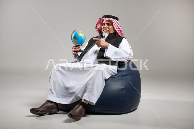 مفهوم البحث والاستكشاف حول العالم, الاشارة إلى مكان معين على الخريطة والنظر إلى أحد القارات, بورتريه لرجل عربي خليجي سعودي كبير بالسن يرتدي الزي السعودي التقليدي والستره السوداء  يجلس على اريكه مريحه من الجلد الفاخر يمسك بيده مجسما للكره الارضيه ويشير اليها، خلفيه رماديه