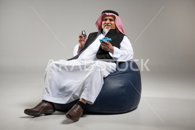 مفهوم القياده في السعوديه، الفرح والسرور بامتلاك سياره حديثه، بورتريه لرجل عربي خليجي سعودي كبير بالسن يرتدي الزي السعودي التقليدي والستره السوداء يجلس على اريكه مريحه من الجلد يمسك بيده مجسم لسياره صغيره ومفتاح القياده، خلفيه رماديه