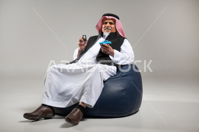 مفهوم القياده في السعوديه، الفرح والسرور بامتلاك سياره حديثه، بورتريه لرجل عربي خليجي سعودي كبير بالسن يرتدي الزي السعودي التقليدي والستره السوداء يجلس على اريكه مريحه من الجلد يمسك بيده مجسم لسياره صغيره ومفتاح القياده، خلفيه رماديه