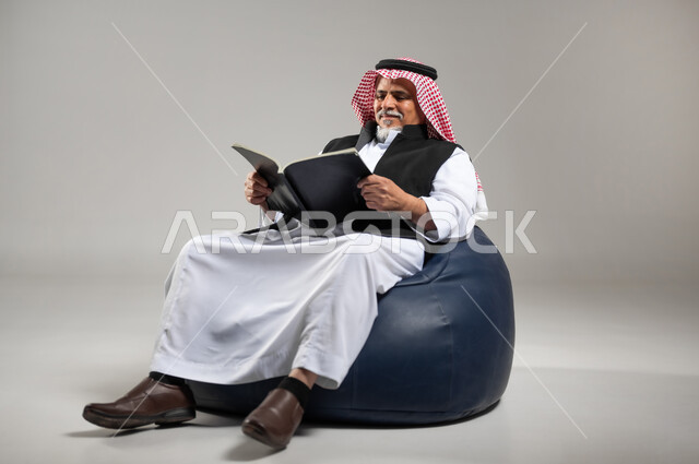 التزود بالعلم والمعرفة، قضاء وقت الفراغ بالتثقيف والتعلم، مفهوم محو الأمية، بورتريه لرجل كبير بالسن عربي سعودي خليجي يرتدي الشماغ والثوب التقليدي يجلس على اريكه مريحه من الجلد يمسك كتابا بيده يقوم بالقراءه، خلفية رماديه
