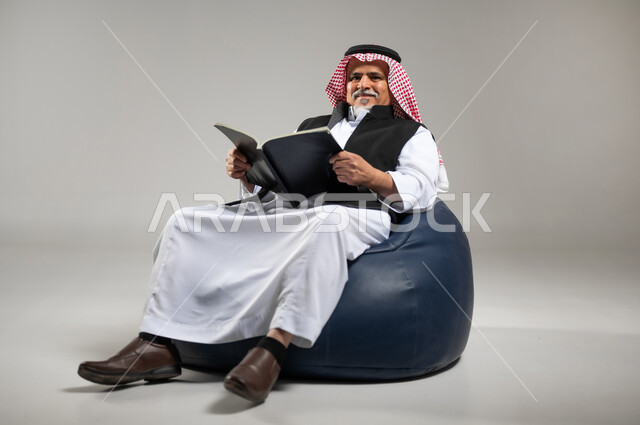 التزود بالعلم والمعرفة، قضاء وقت الفراغ بالتثقيف والتعلم، مفهوم محو الأمية، بورتريه لرجل كبير بالسن عربي سعودي خليجي يرتدي الشماغ والثوب التقليدي يجلس على اريكه مريحه من الجلد يمسك كتابا بيده يقوم بالقراءه، خلفية رماديه