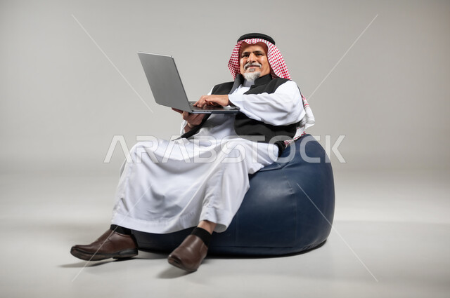 انجاز الاعمال والمهام عن بعد عبر الحاسب المحمول، تصفح الانترنت ووسائل التواصل الاجتماعي، بورتريه  لرجل عربي سعودي خليجي كبير بالسن يرتدي الشماغ والثوب التقليدي والستره السوداء  يجلس على اريكه جلد مريحه يقوم بالعمل على جهاز الاب توب، خلفية رماديه