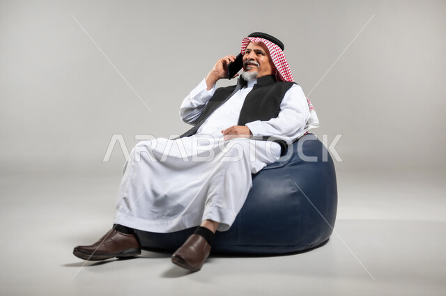 التواصل مع الأصدقاء والاقارب باستخدام الجوال، الرد على الاتصالات والمكالمات، سماع أخبار سعيدة عبر الهاتف المحمول، بورتريه مقرب لرجل عربي سعودي خليجي كبير بالسن يرتدي الشماغ والثوب التقليدي والستره السوداء يجلس على كرسي مريح يقوم بإجراء مكالمة هاتفية، خلفية رماديه