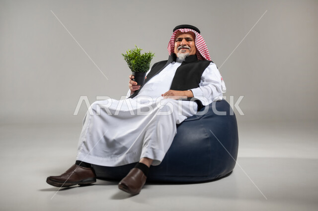 الاهتمام بالشتلات المنزلية, العناية بنباتات الزينة, بورتريه لرجل عربي خليجي سعودي كبير بالسن يرتدي الزي السعودي التقليدي والستره السوداء يجلس على اريكه جلد مريحه يحمل بيده نبته خضراء للزينه, مفهوم الزراعه المنزليه واهميتها في الحد من الانبعاثات الكربونيه، خلفية رمادية