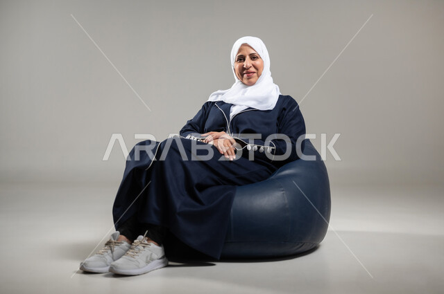 الاستمتاع بقضاء الاوقات على الكراسي المريحه، بورتريه لسيده عربيه خليجيه سعوديه كبيره بالسن مبتسمه ترتدي الحجاب والعباءه السوداء تجلس على اريكه مريحه من الجلد، ايماءات الراحه والاسترخاء، الانتظار ومشاهده التلفاز، خلفيه رماديه