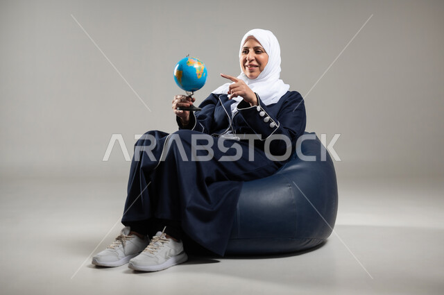 مفهوم البحث والاستكشاف حول العالم, الاشارة إلى مكان معين على الخريطة والنظر إلى أحد القارات, بورتريه لسيده عربيه خليجيه سعوديه كبيره بالسن ترتدي الحجاب والعباءه السوداء تجلس على اريكه مريحه من الجلد الفاخر تمسك بيدها مجسما للكره الارضيه وتشير اليها، خلفيه رماديه