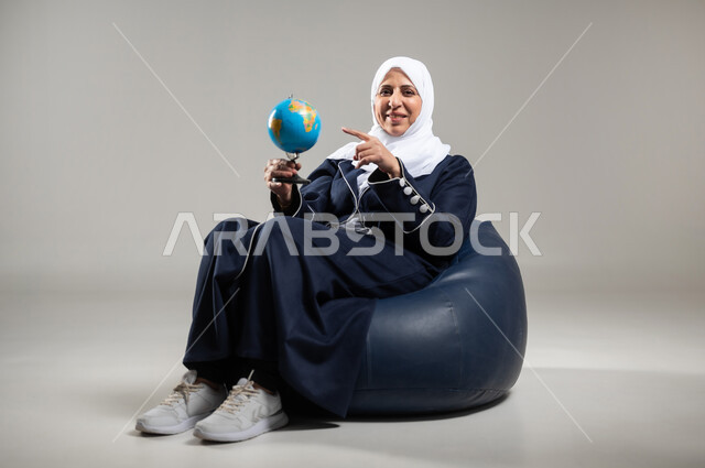 مفهوم البحث والاستكشاف حول العالم, الاشارة إلى مكان معين على الخريطة والنظر إلى أحد القارات, بورتريه لسيده عربيه خليجيه سعوديه كبيره بالسن ترتدي الحجاب والعباءه السوداء تجلس على اريكه مريحه من الجلد الفاخر تمسك بيدها مجسما للكره الارضيه وتشير اليها، خلفيه رماديه