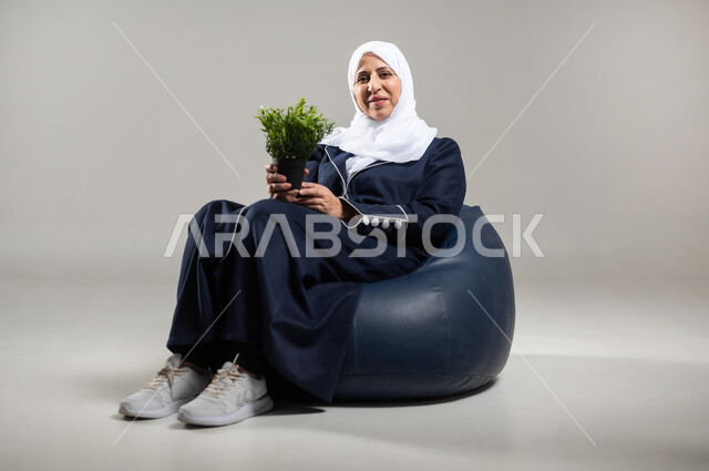 الاهتمام بالشتلات المنزلية, العناية بنباتات الزينة, بورتريه لسيده عربيه خليجيه سعوديه كبيره بالسن ترتدي الحجاب والعباءه السوداء تحمل بيدها نبته خضراء للزينه, مفهوم الزراعه المنزليه واهميتها في الحد من الانبعاثات الكربونيه، خلفية رمادية