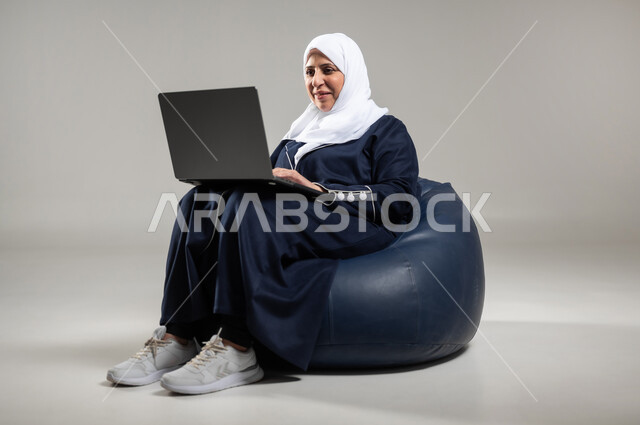 Remote communication and use of social applications using a laptop, portrait of an elderly veiled Saudi Arabian Gulf woman wearing a black abaya sitting on a comfortable leather sofa and working on a laptop, gray background