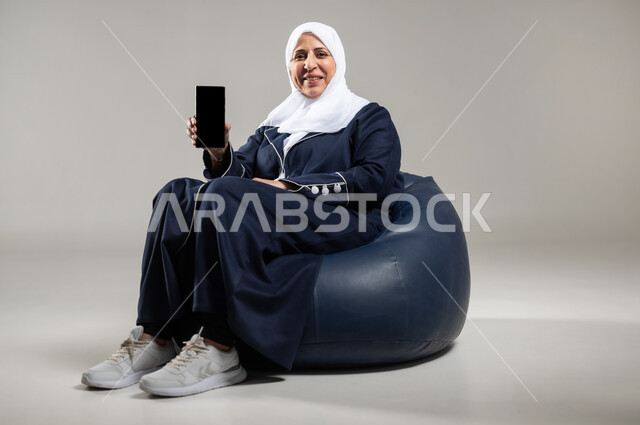 التواصل عن بعد واستخدام التطبيقات الاجتماعيه باستخدام الهاتف المحمول ، بورتريه لسيده عربيه خليجيه سعوديه محجبه كبيره بالسن ترتدي العباءه السوداء تجلس على أريكه مريحه من الجلد وتقوم بعرض الجوال بشاشه سوداء فارغه، خلفيه رماديه