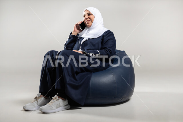 التحدث مع الاقارب والاهل والاصدقاء، بورتريه لسيده عربيه خليجيه سعوديه محجبه كبيره بالسن ترتدي العباءه السوداء تجلس على أريكه مريحه من الجلد وتقوم باجراء مكالمه هاتفيه بايماءات السعاده والسرور، خلفيه رماديه