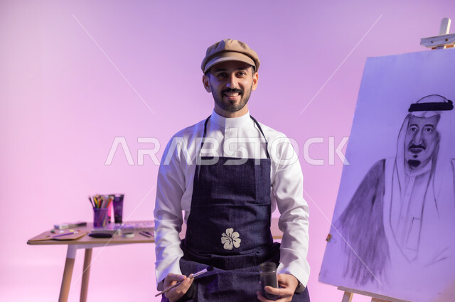 تعابير الفخر والاعتزاز، النظر للكاميرا بإيماءات السعادة، استخدام ريشة الرسم و التلوين، ممارسة مهارات مسلية وممتعة، الجلوس أمام لوحة معالم وعلم المملكة، بورتريه مقرب لرسام عربي خليجي سعودي يرتدي الثوب التقليدي ومريلة الرسم، استذكار اليوم الوطني، خلفية ملونة