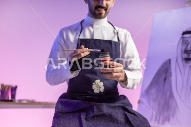 استخدام ريشة الرسم و التلوين، ممارسة مهارات مسلية وممتعة، الجلوس أمام لوحة معالم وعلم المملكة، بورتريه مقرب لرسام عربي خليجي سعودي يرتدي الثوب التقليدي ومريلة الرسم، استذكار اليوم الوطني، خلفية ملونة