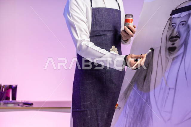 أعياد ومناسبات وطنية، إحياء ذكرى اليوم الوطني السعودي، استخدام ريشة الرسم والتلوين، القيام برسم لوحة معالم وعلم المملكة، التباهي والفخر بالهوية السعودية، حب الوطن والإنتماء إليه، بورتريه مقرب لرسام عربي خليجي سعودي يرتدي الثوب التقليدي ومريلة الرسم، خلفية ملونة