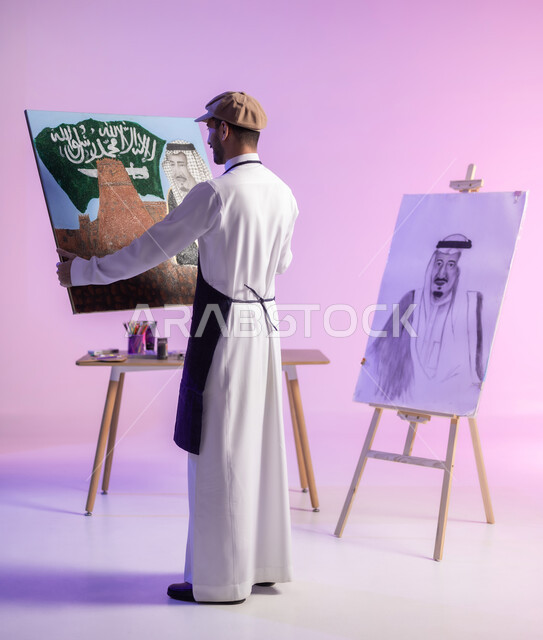 النظر للوحة بإيماءات السعادة، التباهي والفخر بالهوية السعودية، حب الوطن والإنتماء إليه، إحياء ذكرى اليوم الوطني، بورتريه لرسام عربي خليجي سعودي يرتدي الثوب التقليدي ومريلة الرسم يحمل لوحة معالم وعلم المملكة، الجسم بالطول الكامل، خلفية ملونة