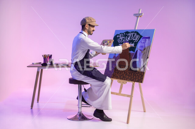 ممارسة هواية الفنون اليدوية، النظر للوحة بإيماءات التأمل والتفكير، استخدام ريشة الرسم والتلوين، الجلوس أمام لوحة معالم وعلم المملكة، بورتريه لرسام عربي خليجي سعودي يرتدي الثوب التقليدي ومريلة الرسم، إحياء ذكرى اليوم الوطني، الجسم بالطول الكامل، خلفية ملونة