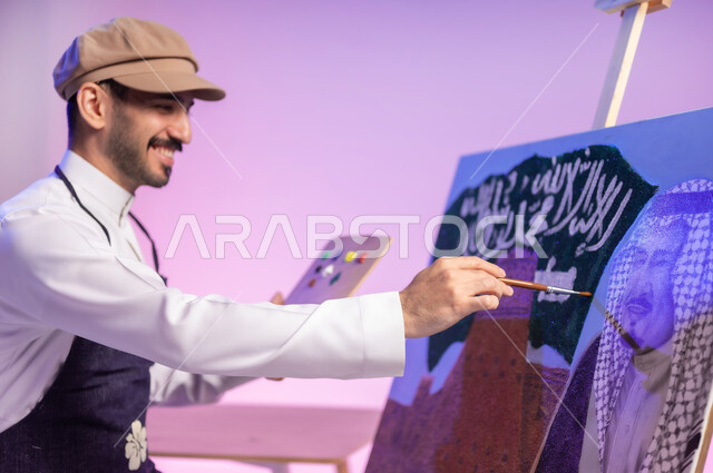 النظر للوحة بإيماءات السعادة والفخر، استخدام ريشة الرسم والتلوين، الجلوس أمام لوحة معالم وعلم المملكة، ممارسة هواية الفنون اليدوية، بورتريه مقرب لرسام عربي خليجي سعودي يرتدي الثوب التقليدي ومريلة الرسم، إحياء ذكرى اليوم الوطني، خلفية ملونة