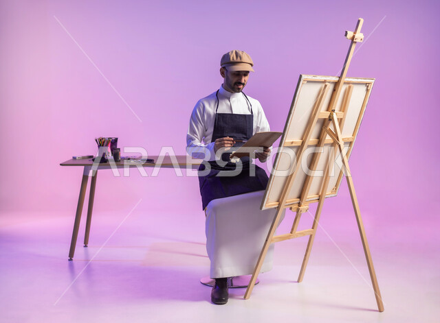 أنشطة ترفيهية فنية ممتعة، استخدام ريشة الرسم والتلوين، ممارسة الهوايات المفضلة، الجلوس أمام اللوحة، بورتريه لرسام عربي خليجي سعودي يرتدي الثوب التقليدي ومريلة الرسم، الجسم بالطول الكامل، خلفية ملونة